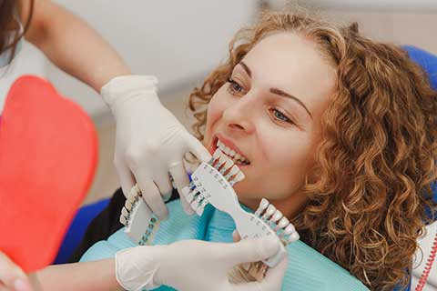 woman sites in a dental chair while the dentist compares which shade of tooth color matches the patient's teeth